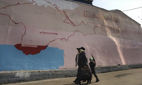 Women and a boy with a toy Kalashnikov rifle walk past an unfinished wall painting depicting a map of Crimean peninsula and a slogan reading 