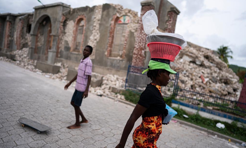 A woman carries goods as she walks in front of a collapsed building in Aquin district, Sud, Haiti, on Aug. 19, 2021. The death toll from a powerful earthquake that struck southwest Haiti on Saturday climbed to 2,189, the Caribbean island's Civil Protection Agency reported on Wednesday.Photo:Xinhua