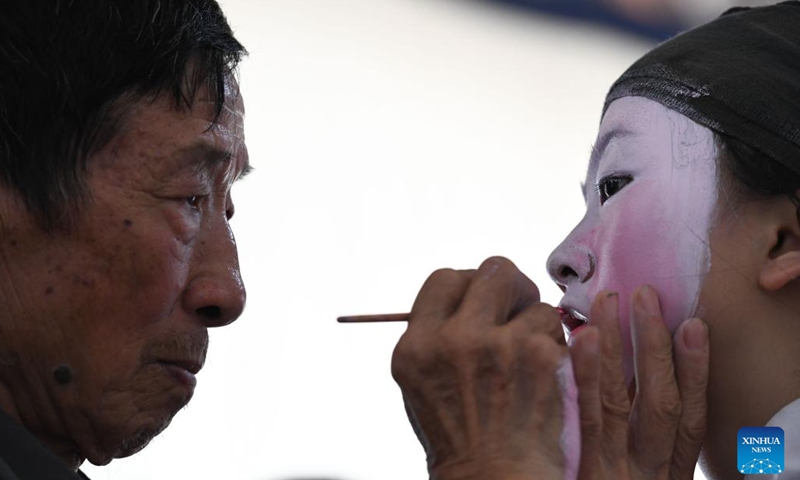 Elderly folk artist Shao Maokai (L) helps 12-year-old Anhui Opera student Wang Yuqin put on makeup at Fuling Village of Jixi County, Xuancheng City, east China's Anhui Province, May 30, 2023. In order to promote Anhui Opera and raise awareness of the traditional performing art, a primary school in Fuling Village has been offering a routine training program since 2003 for its students, hiring veteran folk artists as tutors.(Photo: Xinhua)