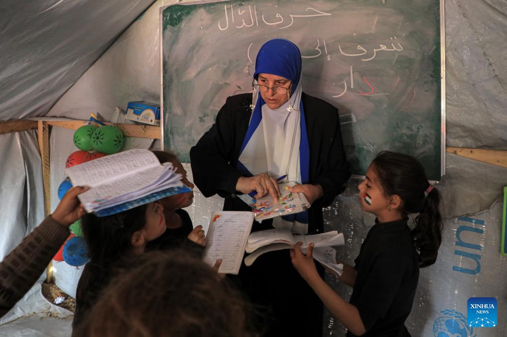 A Palestinian teacher teaches children at a temporary classroom in the southern Gaza Strip city of Rafah, March 27, 2024. The heavy bombardment against Gaza by Israel has resulted in direct hits on 212 schools within the enclave, according to analysis partnered with the United Nations released on Wednesday. Satellite images have shown that at least 53 schools have been completely destroyed since the conflict began on Oct. 7, 2023.(Photo: Xinhua)