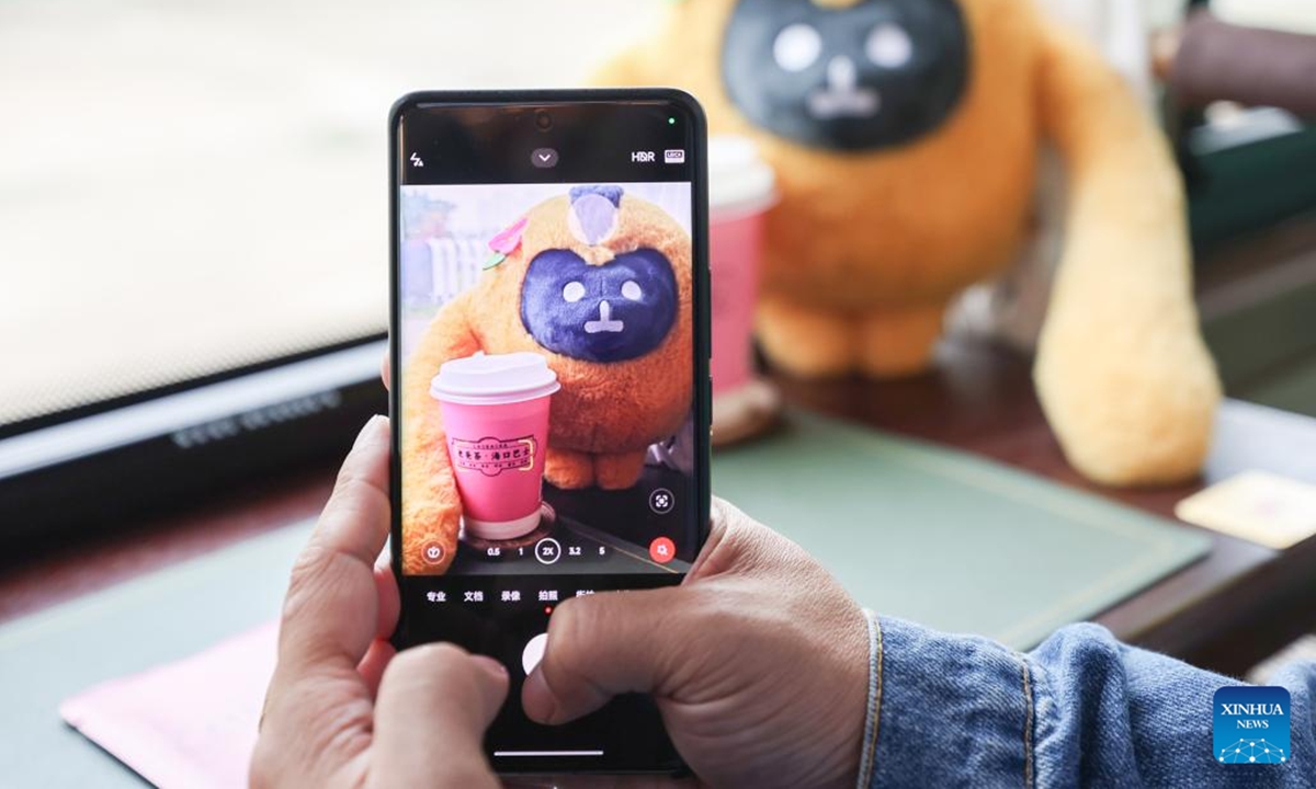 A tourist takes photos of the mascot of the fifth China International Consumer Products Expo (CICPE) on the Laobacha Haikou Bus's CICPE special route in Haikou, south China's Hainan Province, April 6, 2025. Ahead of the upcoming fifth China International Consumer Products Expo (CICPE), the Laobacha Haikou Bus has inaugurated its CICPE special route. Laobacha is a beloved Hainan tradition where locals gather over fragrant tea, savory snacks, and leisurely chat.  (Photo: Xinhua)