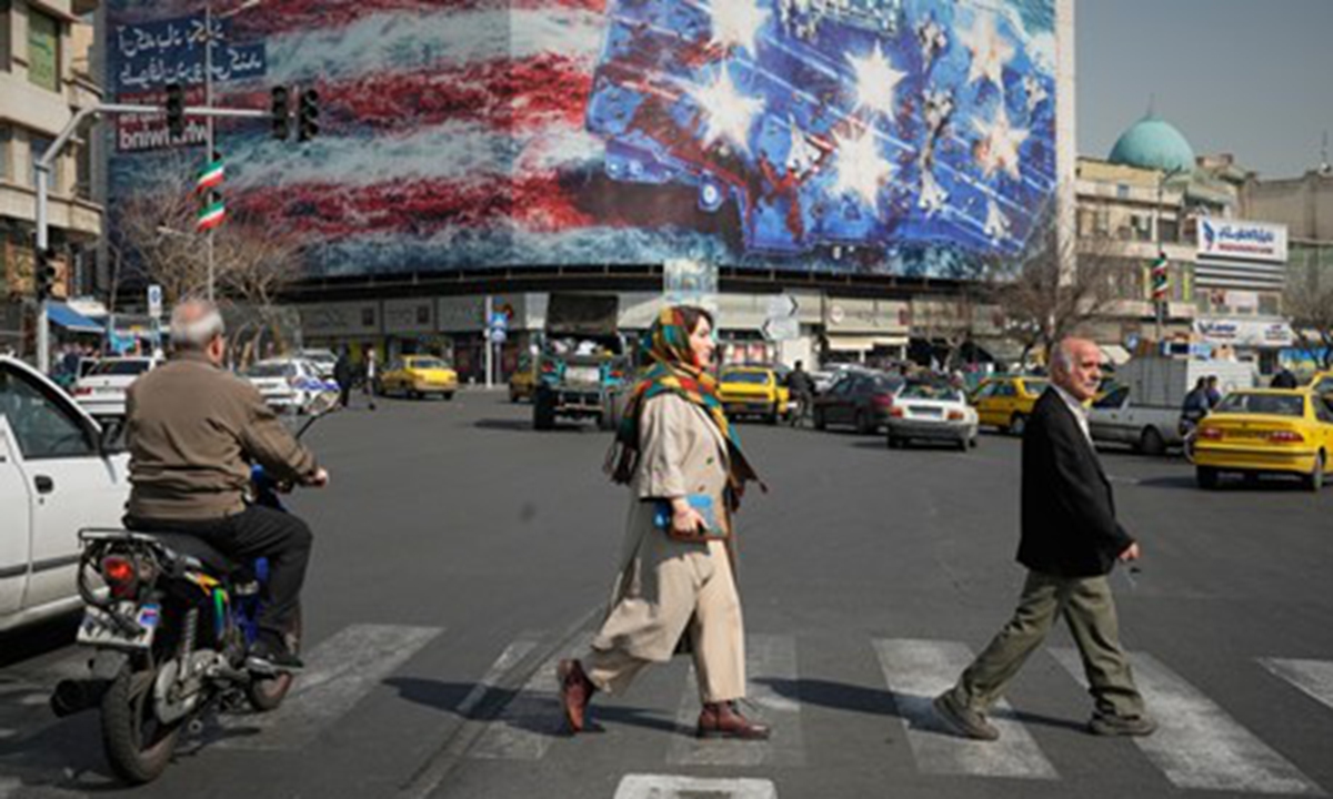 Pedestrians walk past a billboard depicting a U.S. aircraft carrier with damaged fighter jets on its deck at Enqelab-e-Eslami (Islamic Revolution) Square in Tehran, Iran, on February 22, 2026. 