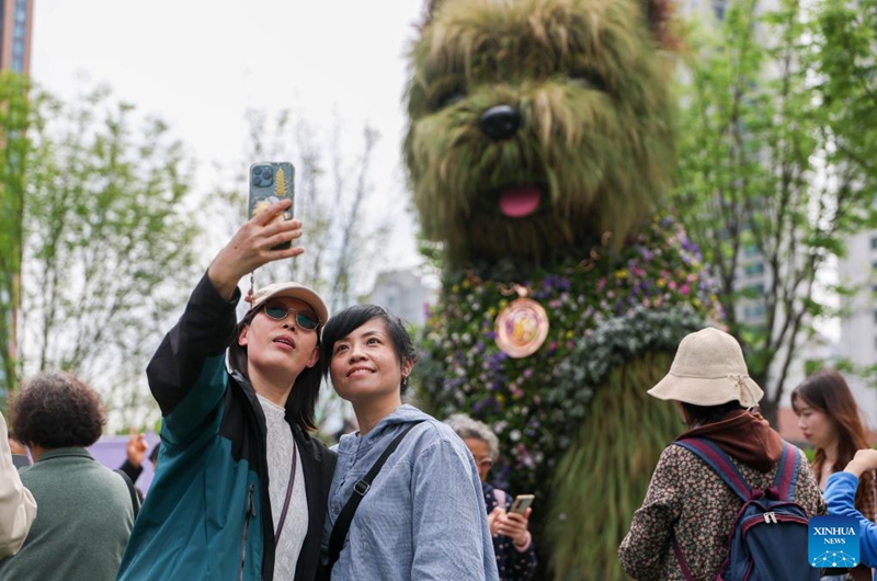 People take a selfie with a floral installation in the shape of a puppy at a flower fair in Jing'an District of Shanghai, east China, April 18, 2026. The 2026 Shanghai International Flower Show opened on Saturday with events in two main venues, 10 sub-venues and multiple exhibition spots across the city. (Xinhua/Wang Xiang)
