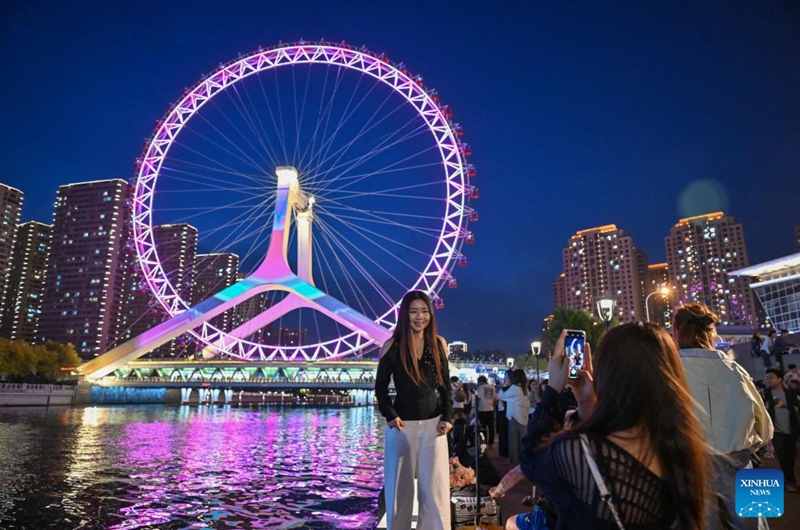 A tourist poses for photos with the Tianjin Eye ferris wheel in Tianjin, north China, April 25, 2026. In recent years, Tianjin has become a popular tourist destination, as the city keeps enriching its nighttime experience by expanding tourism scenarios and diversifying business formats. (Xinhua/Sun Fanyue)