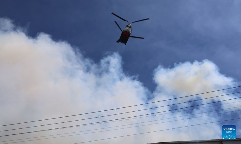 A firefighting helicopter drops water to prevent the fire from a facility in Boyle Heights, Los Angeles, California of the United States, on June 20, 2026. Mayor Karen Bass of the U.S. city of Los Angeles on Saturday declared a local emergency in response to an ongoing warehouse fire that erupted Wednesday and generated heavy smoke, which has spread across the Los Angeles metropolitan area for several days. (Photo by Zeng Hui/Xinhua)

