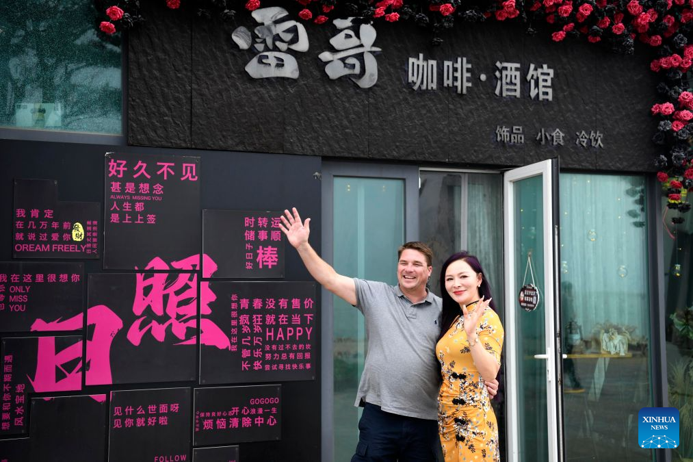 Ryan Norman and his wife Zhang Lei wave hands outside their cafe in Rizhao, east China's Shandong Province, Sept. 24, 2026. (Xinhua/Guo Xulei)