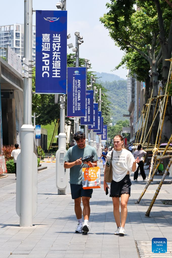 Foreign tourists walk past themed banners on a street in Huaqiangbei of Shenzhen, south China's Guangdong Province, Aug. 10, 2026. With themed bulletin boards, outdoor posts, decorations and installations, Shenzhen marked on Monday the 100-day countdown to the 33rd Asia-Pacific Economic Cooperation (APEC) Economic Leaders' Meeting. (Xinhua/Liang Xu)