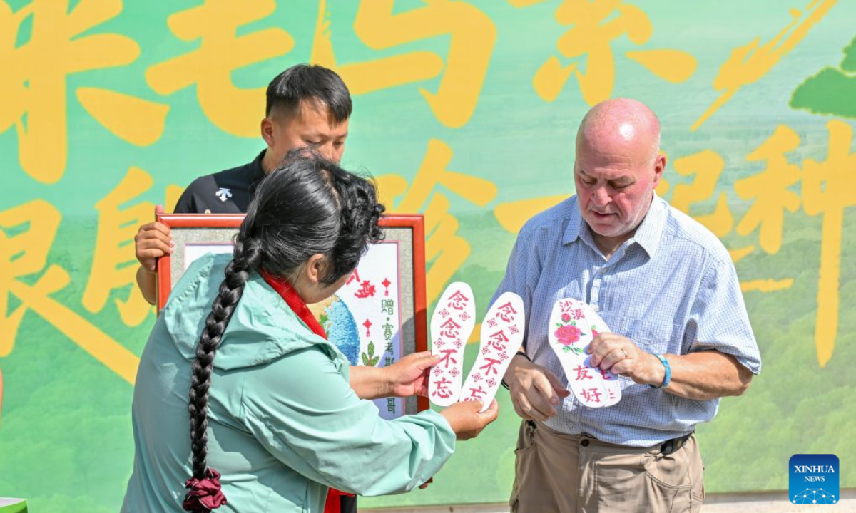 Yin Yuzhen (L) presents Ronald Sakolsky with cross-stitch embroidery and shoe insoles in the Maowusu Sandy Land in north China's Inner Mongolia Autonomous Region, Aug. 25, 2026. Twenty-six years had passed before Yin Yuzhen, a national-honored sand-control villager in China's Inner Mongolia, finally sat down to have a reunion feast with her American friend Ronald Sakolsky.
The story behind the feast began in 1999, when Sakolsky taught English at a school in central China's Henan Province under a teachers' exchange program between the two countries.
By then, Yin had spent more than a decade battling desertification that had severely threatened her livelihood, even half burying her house during sandstorms.
After watching a television news report featuring Yin's tireless work, he was deeply moved and raised 5,000 U.S. dollars to help Yin in her afforestation efforts against desertification.
The saplings bought and planted with that donation have since grown into a forest of more than 50,000 towering trees.
As Sakolsky left Yin's home after their reunion in Maowusu Sandy Land on Tuesday, Yin presented him with gifts she had long prepared. (Xinhua/Sun Ruibo)