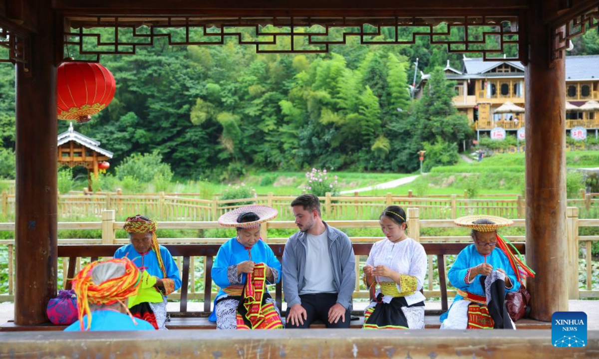 A foreign tourist looks on as local women make Tiaohua cross-stitch embroidery, a traditional handicraft passed down through generations by women of Huayao, a branch of Yao ethnic group, at Huxingshan Yao Township in Longhui County, Shaoyang City, central China's Hunan Province, Aug. 4, 2026. During midsummer, Huxingshan Yao Township embraces a tourism boom thanks to its alpine climate, rich intangible cultural heritage and folk traditions. (Photo by Zeng Yong/Xinhua)