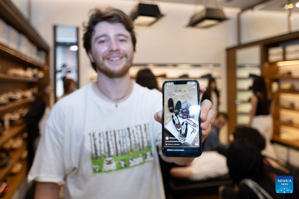 Maksim Ermolaev, a tourist from Russia, shows shoes from a Chinese brand that he has seen on social media at a shop in Shanghai, east China, July 22, 2026. The foreign visitors entering Shanghai increased 22 percent year on year in the first half of 2026, thanks to the country's expanding visa exemption policies. Chinese designer products have emerged as a new driver of inbound consumption. Rather than buying only traditional Chinese specialties such as tea and silk, overseas visitors are increasingly following shopping tips on social media to seek out bags, footwear, clothing and other products by Chinese designers. (Xinhua/Wang Xiang)