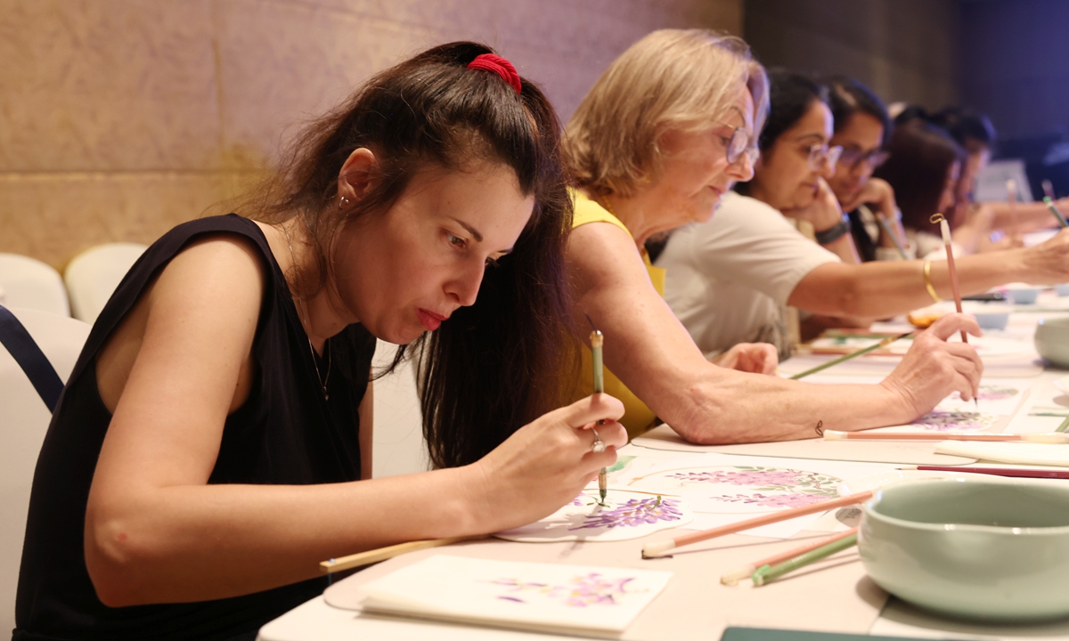 Foreign female participants concentrate on creating ink paintings on round fans during the hands-on Chinese painting experience session of the event.