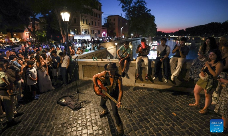 People watch an artist performing along the Tiber River in Rome, Italy, July 12, 2026. The 24th edition of Along the Tiber Rome event is held here along the banks of the Tiber and will run until the end of August. It has become a popular summer event featuring culture, music and gastronomy. (Xinhua/Wang Kaiyan)