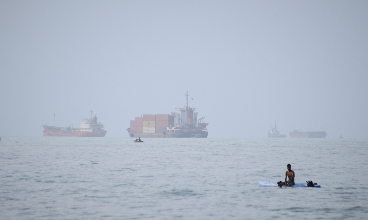 A man sits on a surfboard as a container ship and other commercial vessels appear anchored in the Strait of Hormuz off Bandar Abbas, Iran, July 27, 2026. Photo： VCG