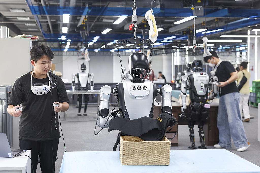 A trainer guides a robot in a clothes-folding exercise at a humanoid robot data training center in Beijing's Shijingshan District, on August 10, 2026. The center, which houses more than 100 humanoid robots, conducts large-scale data processing and model training across 10 real-world application scenarios, including home care, special operations, and new retail, building end-to-end service capabilities. Photo: VCG