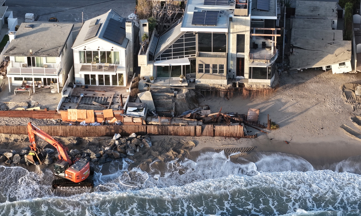 In an aerial view, a Pacific Ocean wave washes up near a worker using heavy machinery to protect damaged beachfront homes with boulders on September 14, 2026, in Dana Point, California.