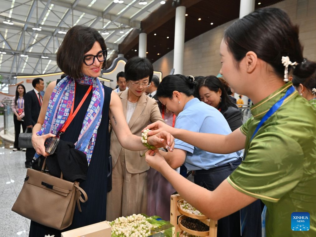 A staff member helps a foreign participant of the 21st Century Maritime Silk Road Local Government Cooperation and International Friendship Cities Dialogue put on a bracelet made of jasmine flowers at a Fujian intangible cultural heritage exhibition in Fuzhou, southeast China's Fujian Province, Oct. 17, 2026. (Xinhua/Jiang Kehong)
