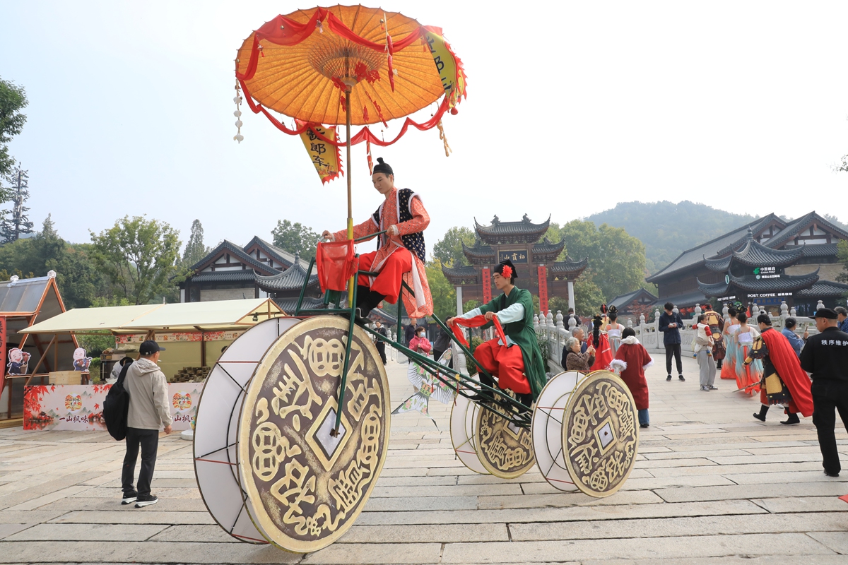 Two performers in ancient-style costumes ride a giant coin tricycle as part of a roaming folk show at the Qixia Mountain scenic area in Nanjing, East China's Jiangsu Province, on October 21, 2026. The scenic area, a rich historical and cultural heritage, hosts a rich lineup of cultural performances for tourists as autumn unfolds. Photo: VCG